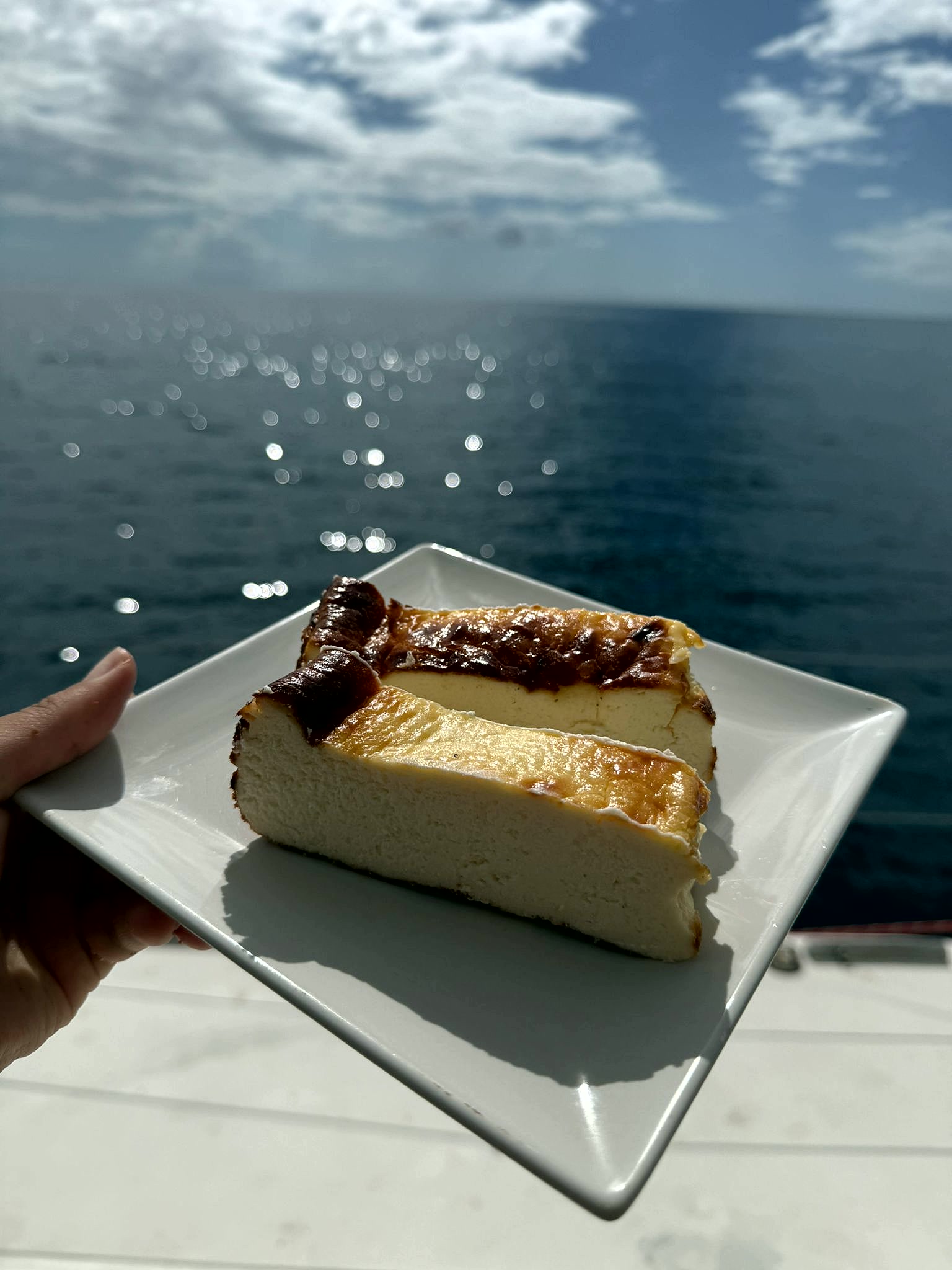 A slice of deeply caramelised Basque burnt cheesecake on a white plate held over the Caribbean sea